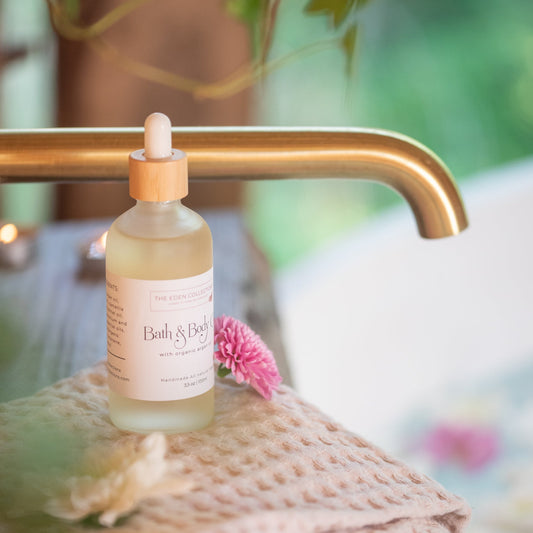 All natural bath and body oil scented with essential oils, enriched with argan oil, poured into a glass container. Lying on a towel among flowers in a modern bathroom. Handmade by The Eden Collections.