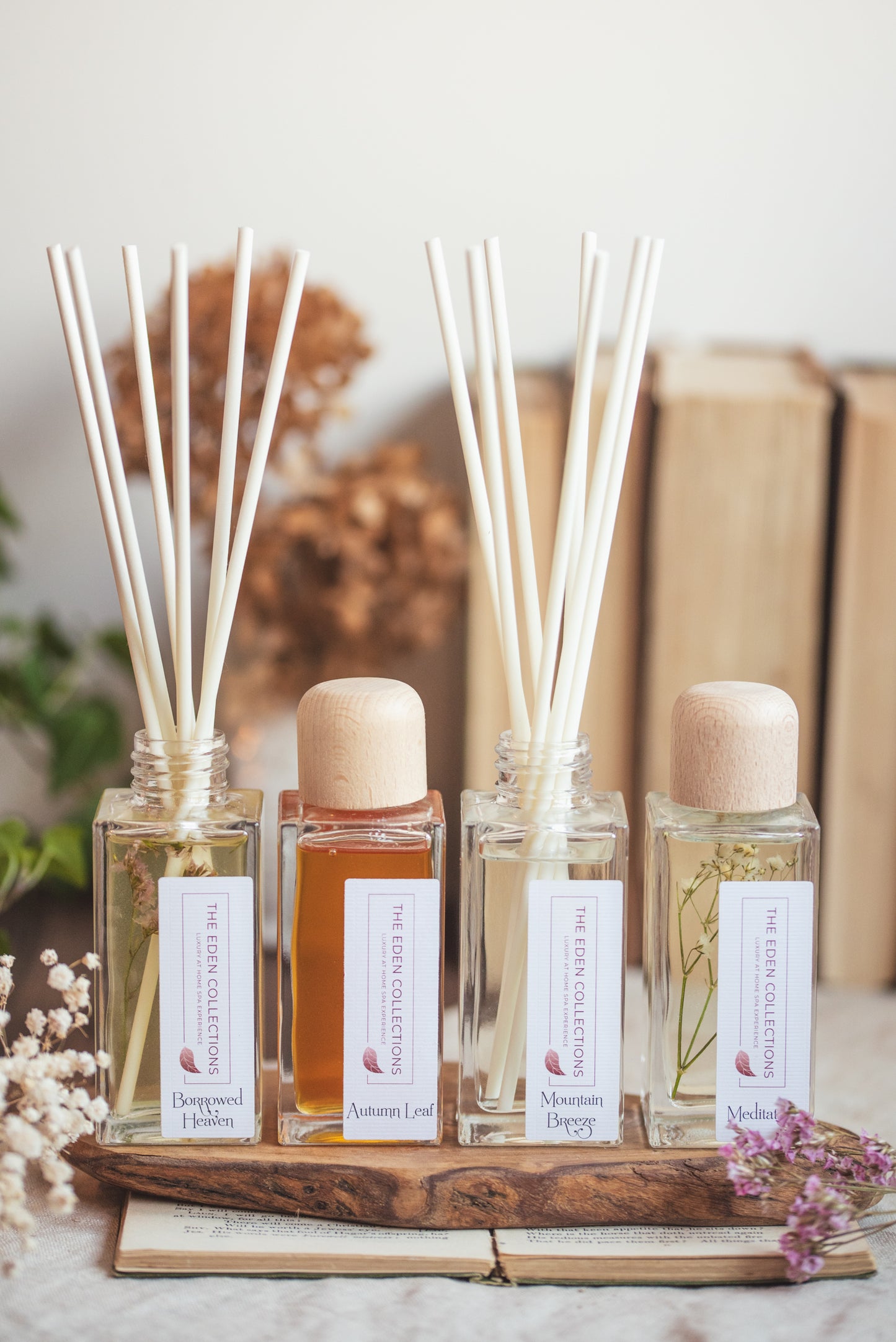 Four elegant, aromatherapy reed diffusers with essential oils, botanicals and fibre reeds. They are showcased together on a wooden tray, with dried flowers and a book.