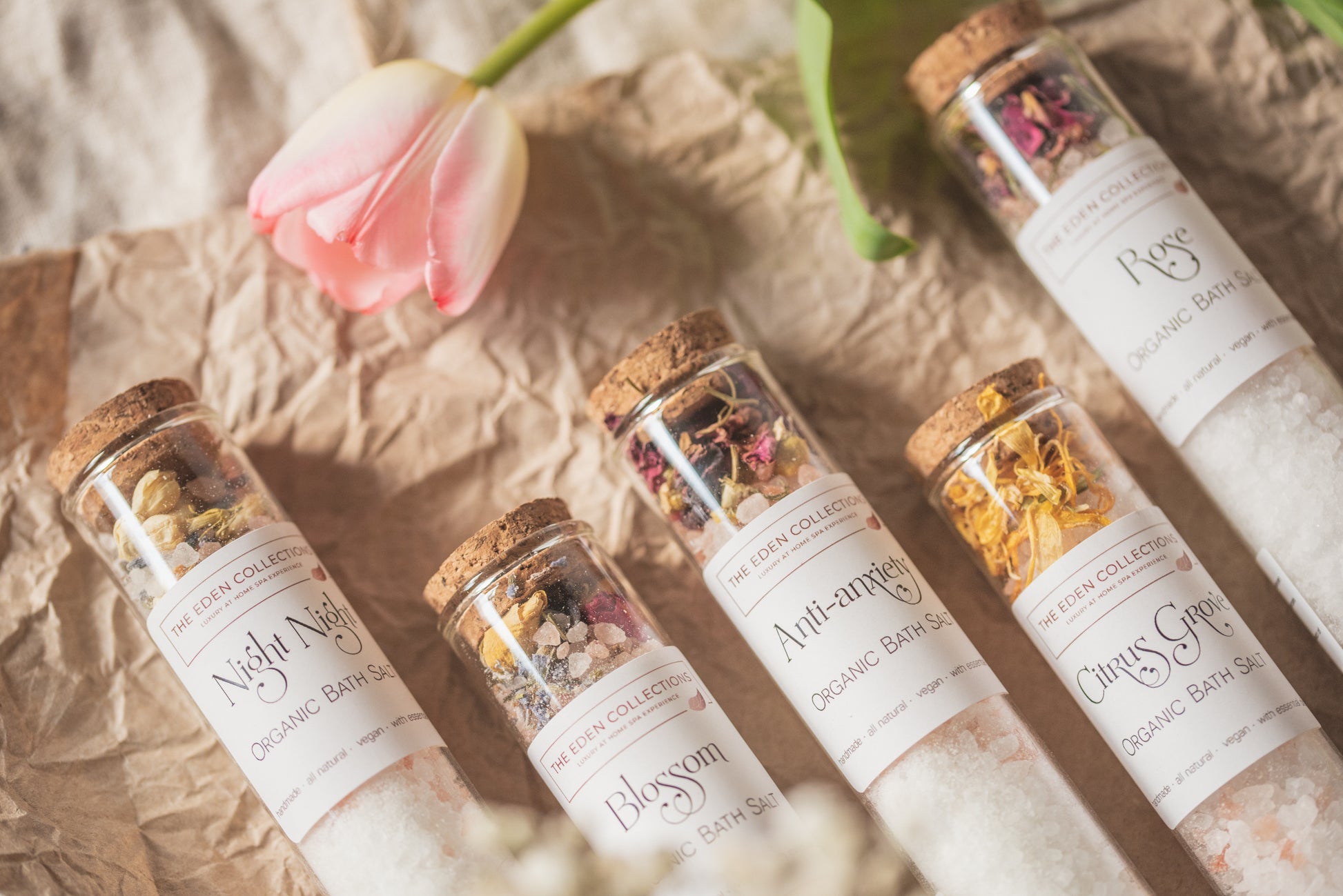 The Eden Collections' organic bath salt shots with essential oils, in glass tube with cork lid and dried botanicals are displayed alongside with a pink tulip. Handmade in Wales.