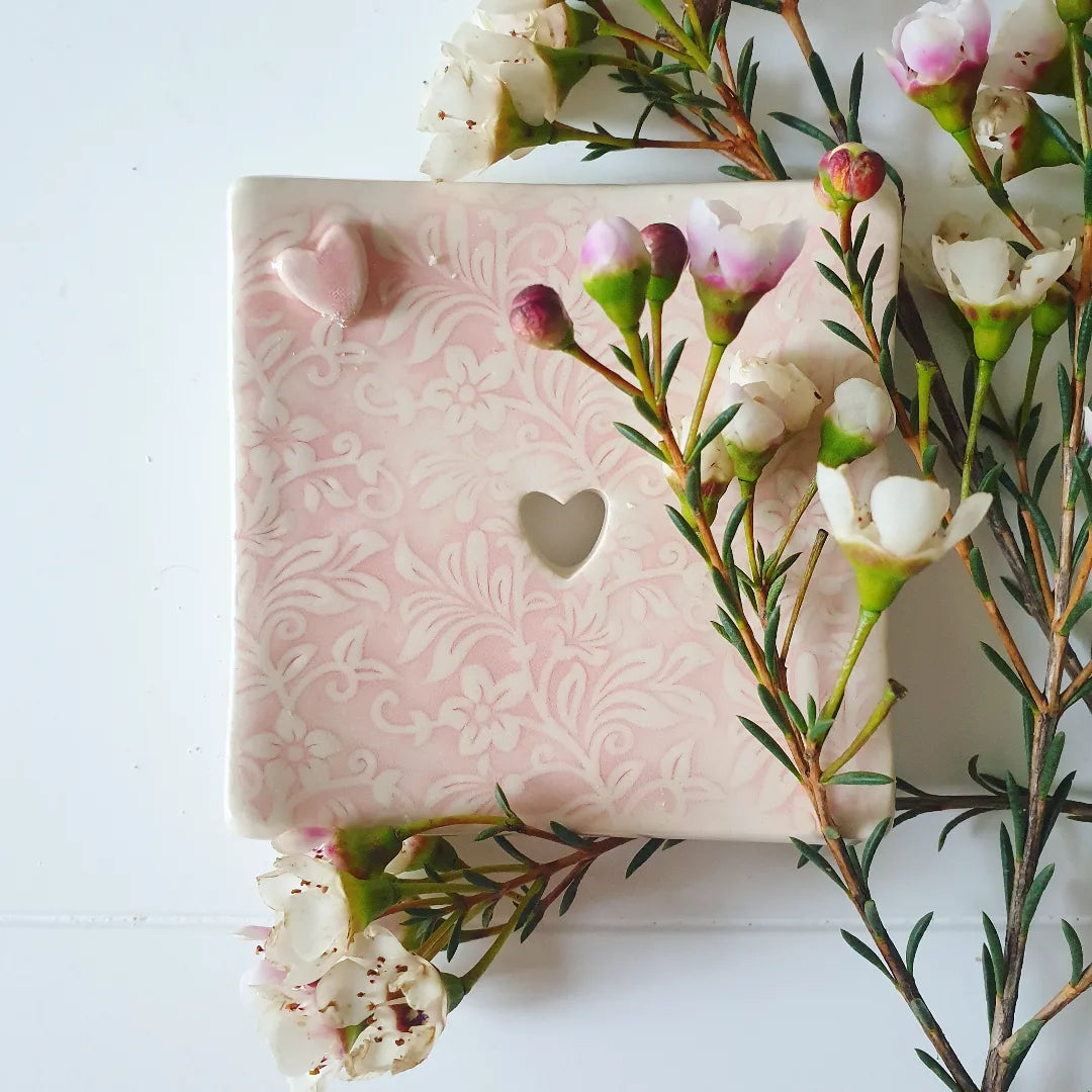 The Eden Collections beautiful floral embossed pink square ceramic soap dish displayed alongside flowers.